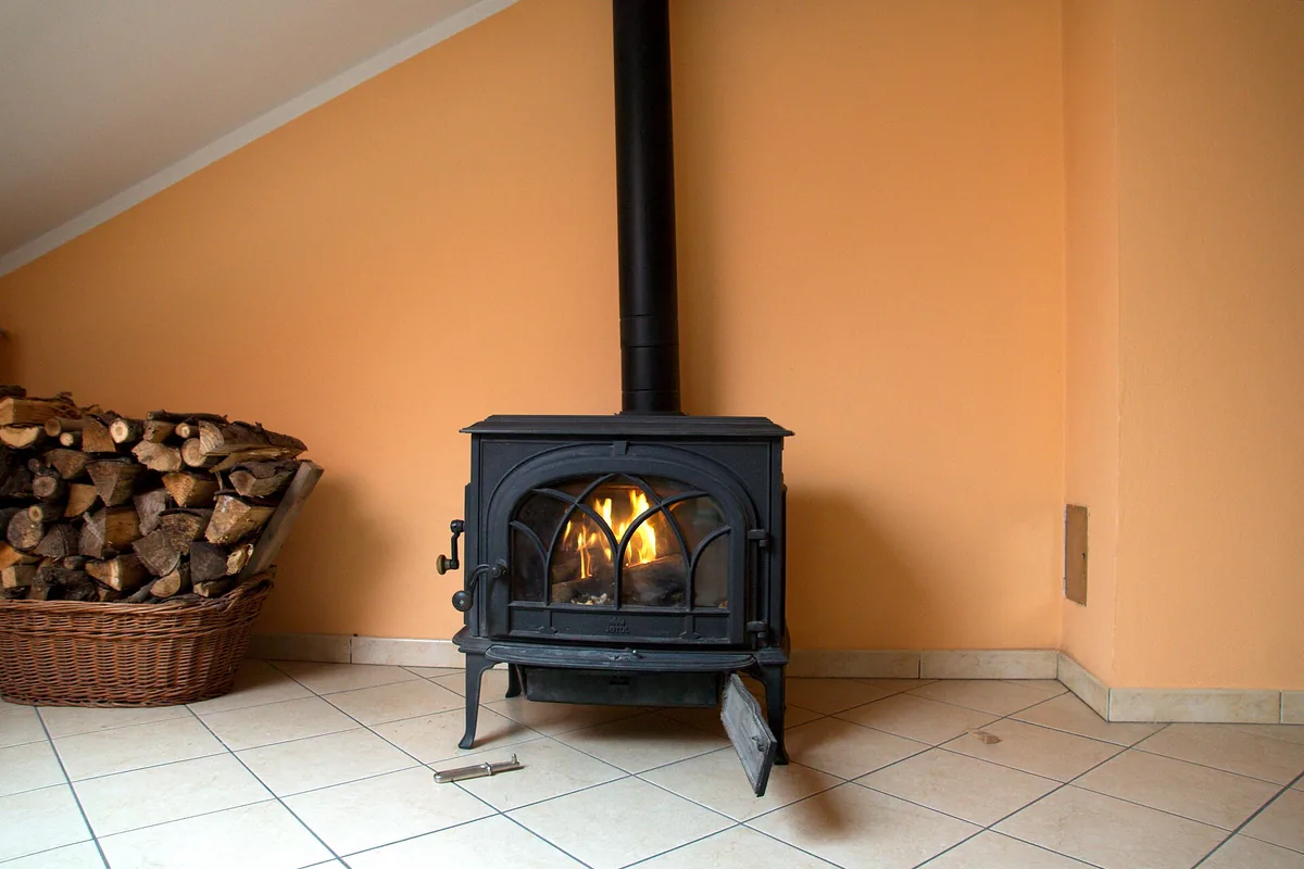 A cast iron wood burning stove with a fire lit behind its glass door and split firewood stacked beside it