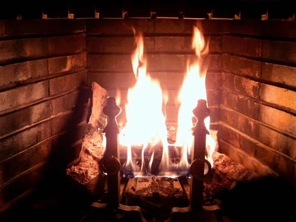 A wood fire burning in a brick fireplace firebox behind iron andirons