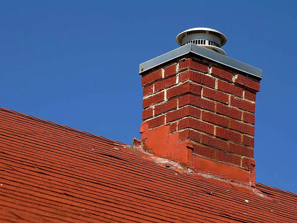 A brick chimney topped with a metal rain cap on a red tile roof